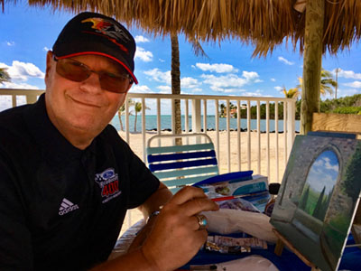 Artist Gilbert Lessard painting one of his art pieces while sitting on a chair on the beach